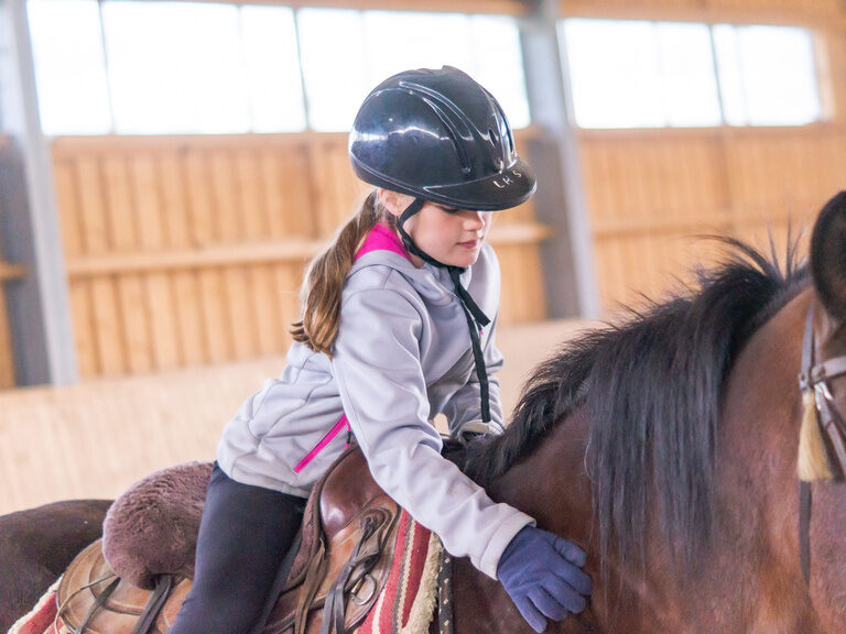 Mädchen sitzt auf Pferd beim Reitunterricht und streichelt den Hals