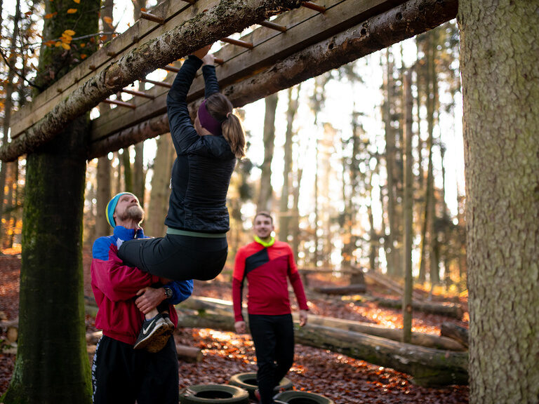 2 Männer und eine Frau beim Outdoor Sport, die Frau hängt an einer Leiter während ein Mann sie hält