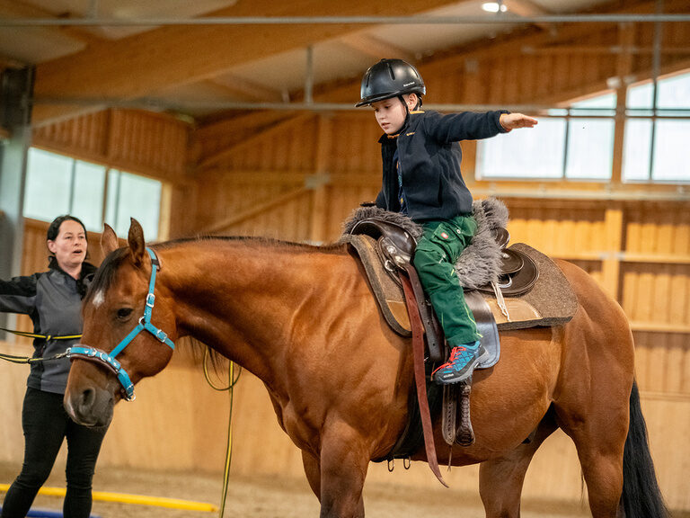 Kind sitzt auf Pferd und streckt die Arme aus während eine Frau daneben die Longe in der Hand hält