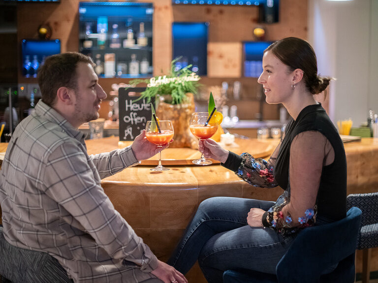 Pärchen sitzt schick gekleidet an der Bar und stößt mit einem Getränk an
