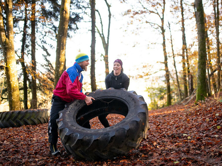 Frau hebt im Wald einen Traktorreifen auf, Mann leistet  Hilfestellung